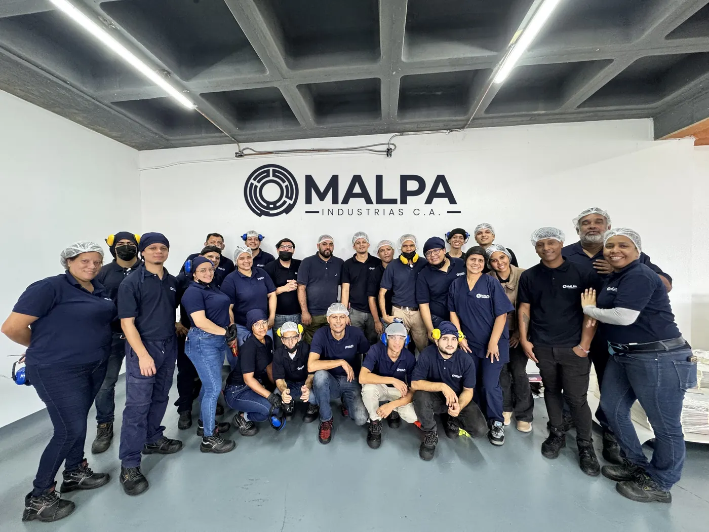 El equipo de planta de Malpa Industrias, con uniforme azul marino, gorro y protectores auditivos, frente a la pared con el logo de la empresa