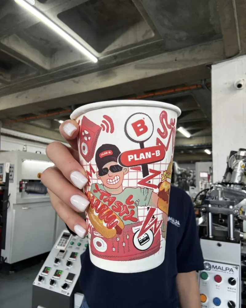 Vaso Plan-B con ilustración de personaje, sostenido en la planta de MALPA